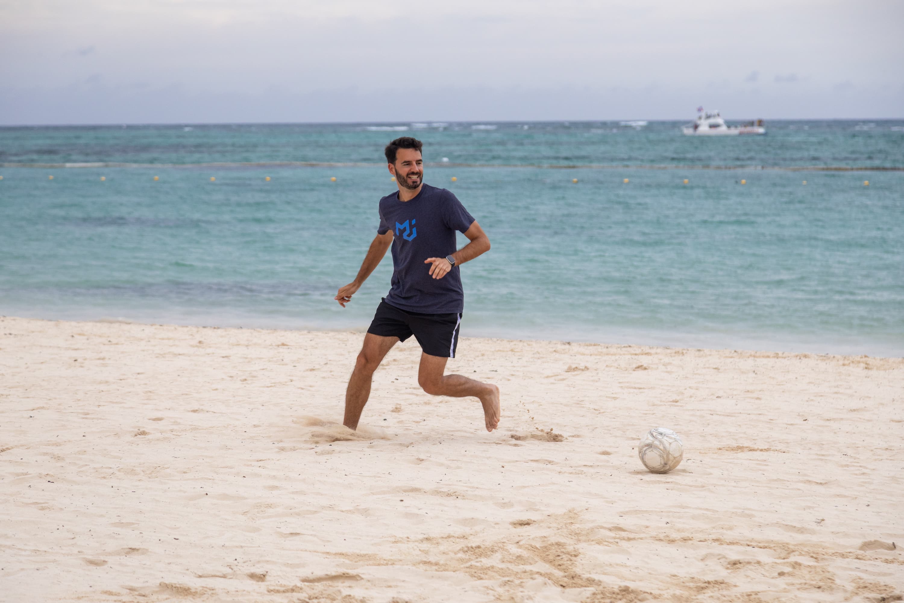 A man with short dark hair and a blue t-shirt prepares to kick a football on a white sand beach.